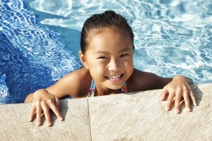 A little girl, about seven, is holding on to the edge of a pool and smiling.