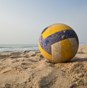 A yellow, white, and blue volleyball sits on the sand with the ocean in the background.