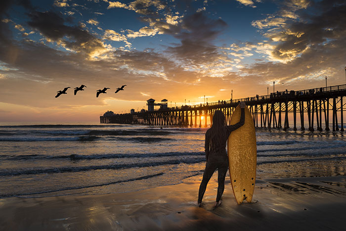 Surfer with long hair in wetsuit standing facing Oceanside Pier with surfboard at sunset